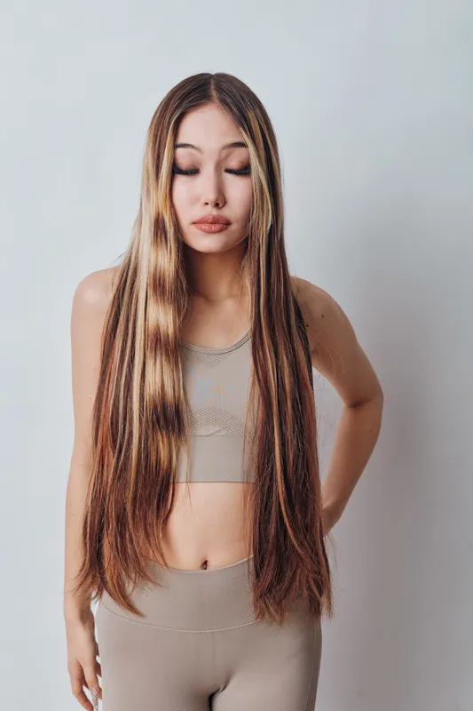 Woman with long braided hair and closed eyes stands in front of plain wall wearing beige crop top and matching pants in casual studio portrait with natural light.