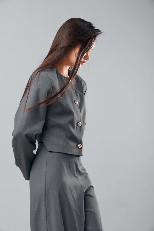 Woman in gray suit standing against gray background with hands behind back looking down. Businesswoman wearing formal outfit, long hair, and buttons on jacket in studio light.