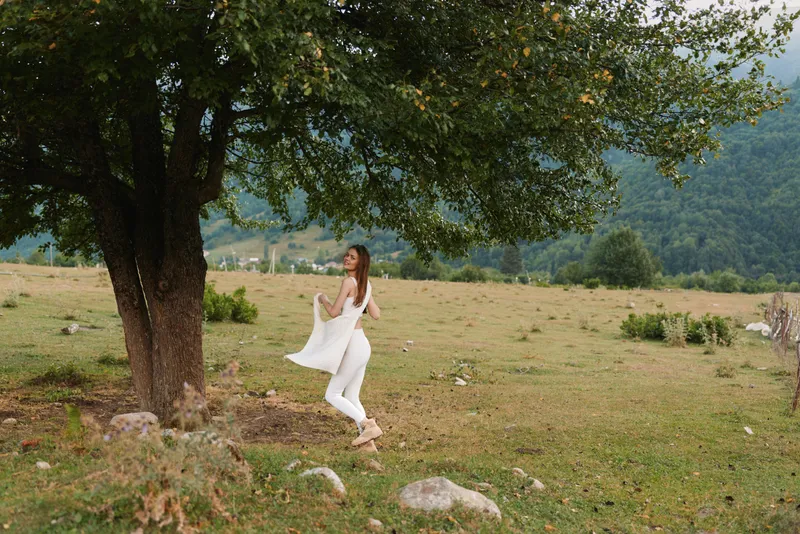Woman in white dress dances beside a tree in an open field, daytime natural scene, playful lifestyle, serene mood, soft sunlight.