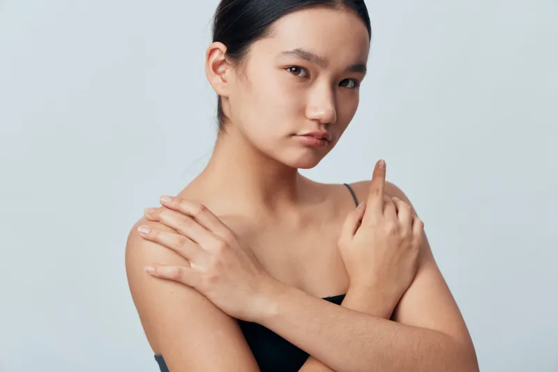 Portrait of a young woman with natural skin posing against a light background, arms crossed over chest, looking calmly at camera with minimal makeup and sleek hair.
