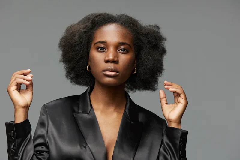 Confident Black woman with natural textured hair in a sleek black blazer posing against a gray background, showcasing empowerment and elegance