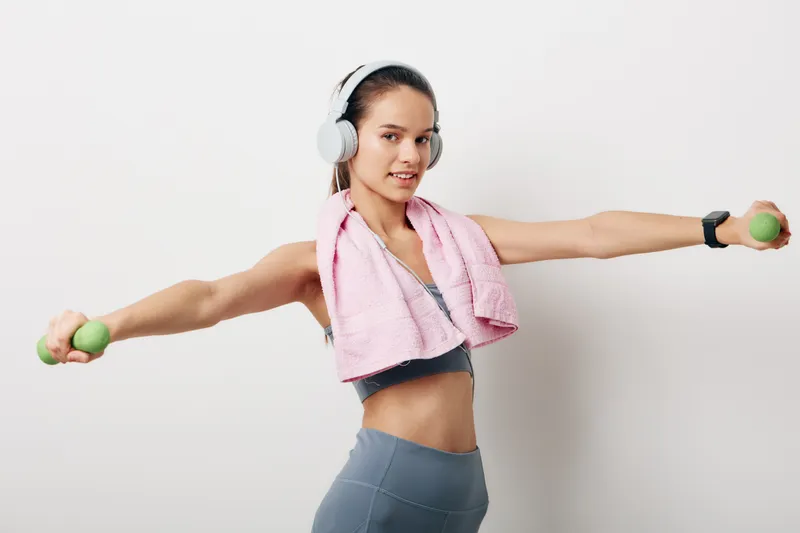 Young woman fitness workout in gray activewear with a pink towel, holding light green dumbbells with arms extended, wearing white headphones and a smartwatch in a bright studio.