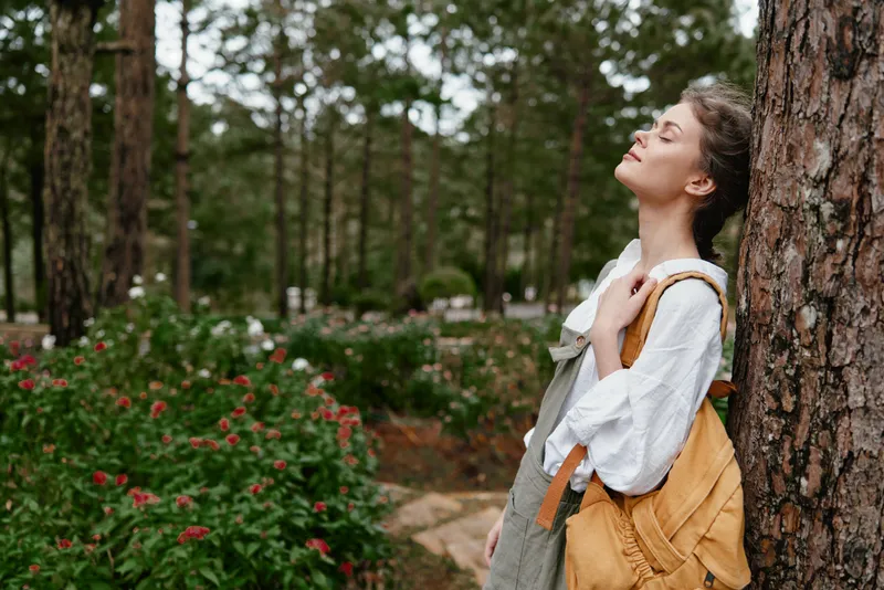 Young woman enjoying nature outdoors with a backpack, wearing casual clothing, set against a serene green forest background in warm tones