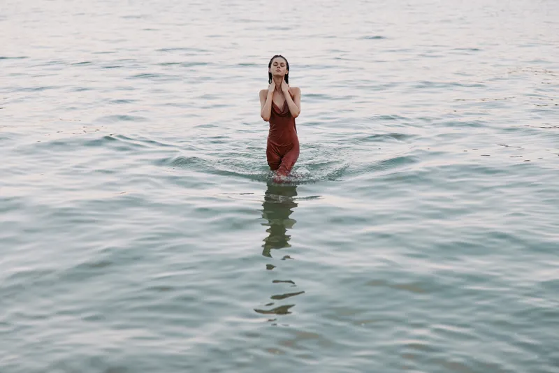 A woman stands waist deep in calm sea water, wearing a red dress, hands raised near her face, creating a serene coastal moment under soft light.