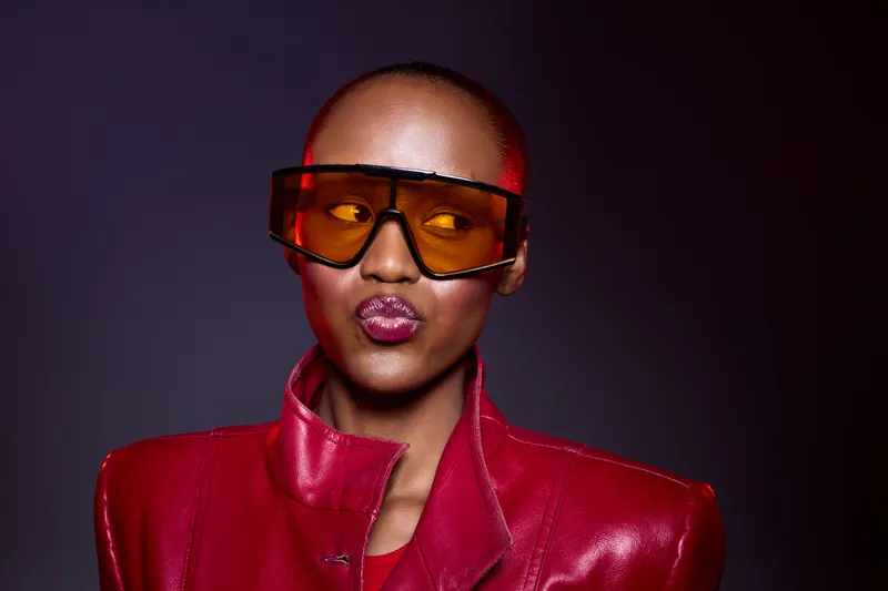 Portrait of confident African woman with dark skin wearing large orange sunglasses and red leather jacket. Studio shot on dark background. Concept of fashion, style, and empowerment.