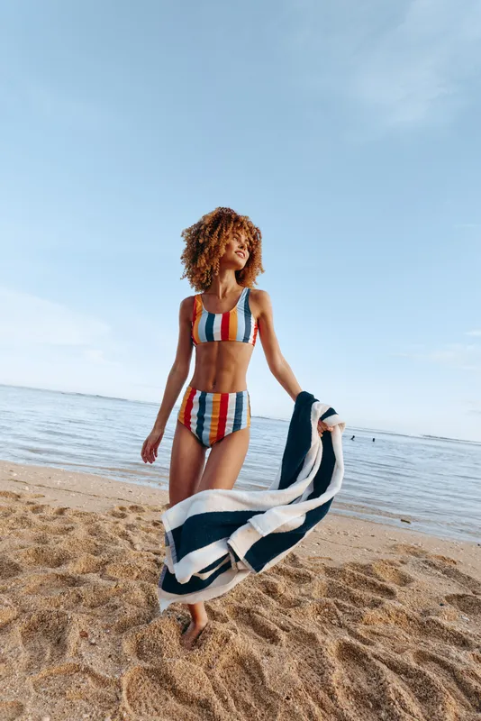 Cheerful woman on the beach in a striped bikini walking along the shoreline with a towel, bright daylight, relaxed pose, blue water, soft breeze, natural colors, sandy background and warmth