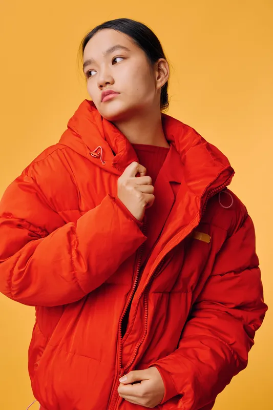 Young woman wearing red jacket posing against orange background, looking sideways with thoughtful expression, fashion portrait with bright outerwear and casual style.