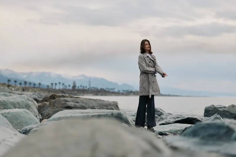 Woman in long coat stands on rocky beach near calm sea under cloudy sky. Winter landscape with distant mountains and palm trees creates serene and contemplative mood.