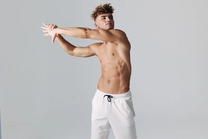 Young man stretching arms and torso for fitness and flexibility in white pants on plain background. Athletic male warming up for workout or sports training session.