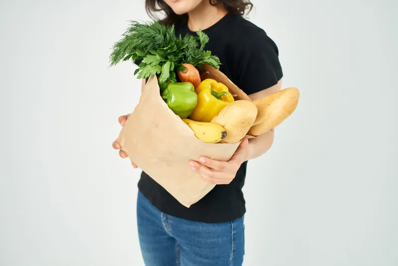 Крупным планом женщина держит в руках пакет с продуктами, в котором находятся овощи, ассоциирующиеся со здоровым питанием