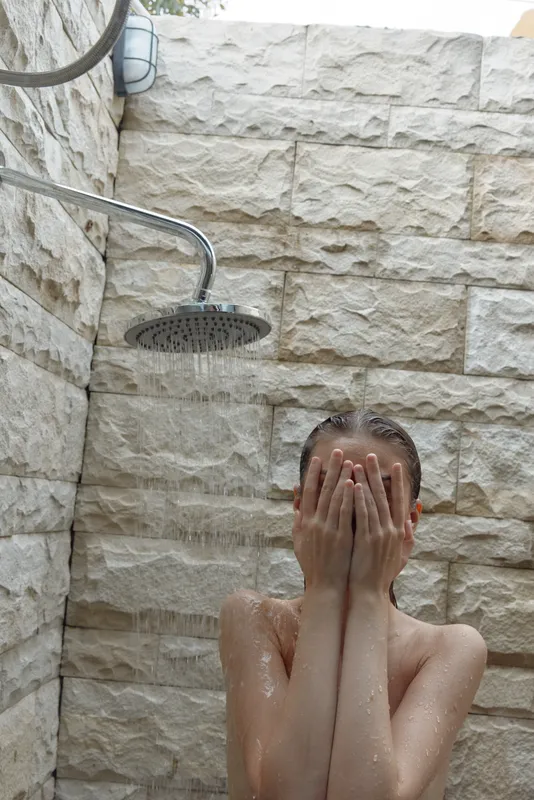 Woman covering face under outdoor shower with stone wall background, natural bathing moment, fresh water droplets, wellness and privacy concept.