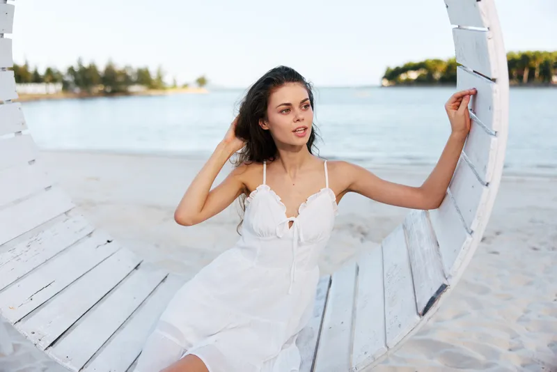A graceful portrait of a young woman in a white sundress seated on a wooden swing by a calm seaside, capturing soft light, relaxed mood, and timeless summer charm.
