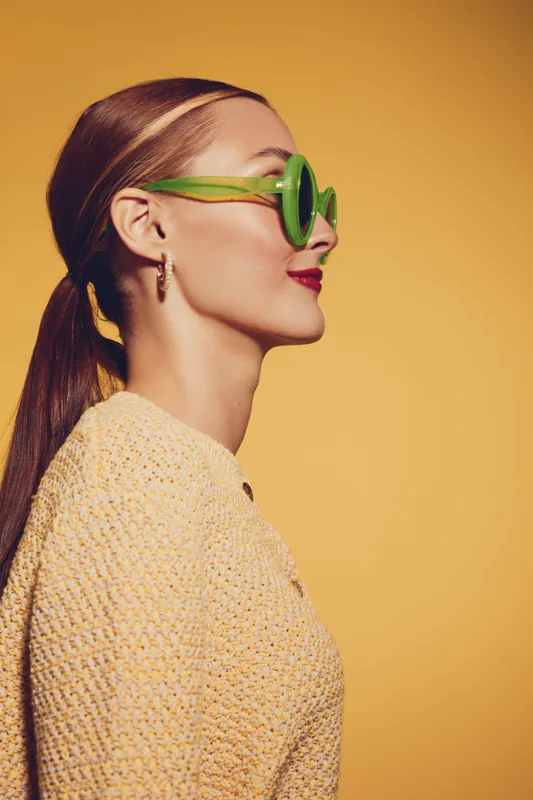 Young woman with green sunglasses and red lipstick poses in profile against yellow background. She wears a textured yellow sweater and has her hair tied back in a ponytail.
