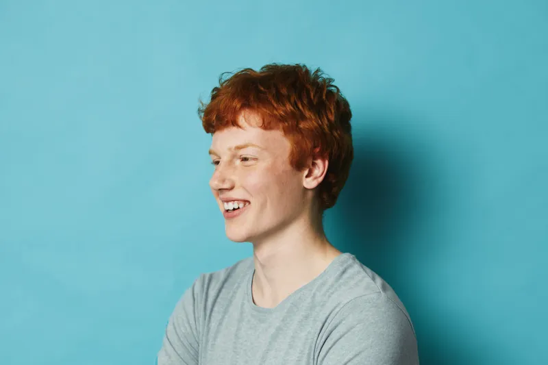 Young man with red hair smiling and looking away on blue background in casual shirt, happy expression and natural light for portrait and lifestyle.
