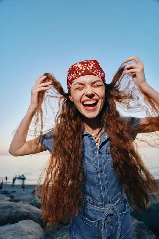 Lifestyle woman with long red hair in modern boho-western denim street style, wearing a colorful bandana, laughing joyfully outdoors with film color tones.