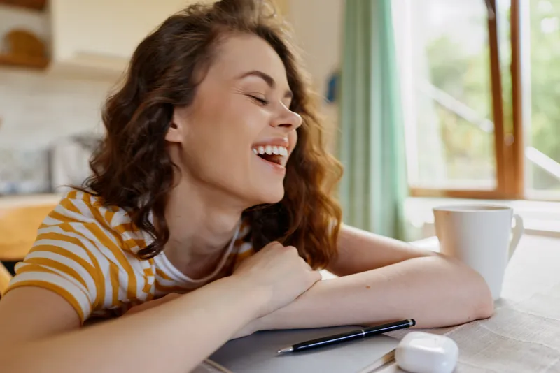 Cheerful woman in a striped tee rests at a sunny kitchen desk, smiles beside a white mug, pen and paper, embodying relaxed home lifestyle and creative flow