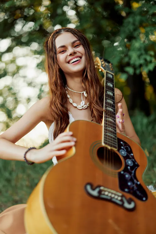 Молодая женщина в стиле хиппи с гитарой в руках сладко улыбается в камеру во время поездки на природу, образ жизни в гармонии