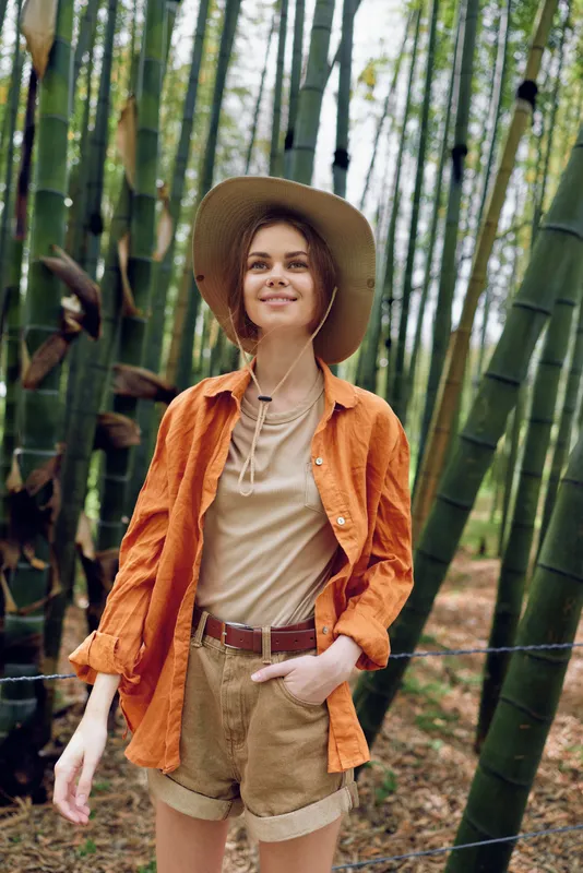 Woman in hat with a warm smile standing among tall bamboo stalks in a dense forest, wearing orange shirt and shorts, casual outdoors portrait capturing nature, travel and peaceful exploration.