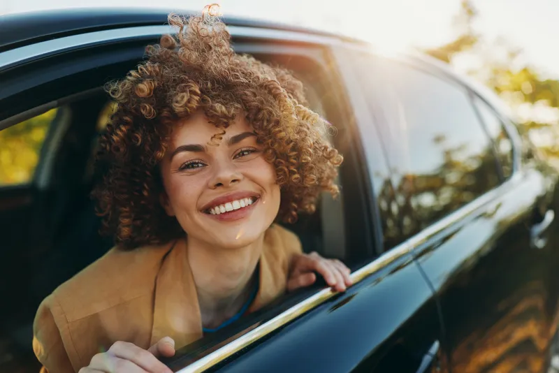 Smiling woman with curly hair leaning out of a car window, sunlit moment, casual fashion, carefree vibe, lifestyle portrait with retro film color and warm tones on social media