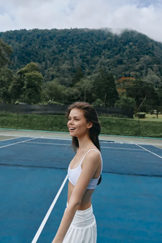 Smiling young woman in sporty outfit playing tennis on outdoor court surrounded by nature, showcasing healthy lifestyle and athleticism
