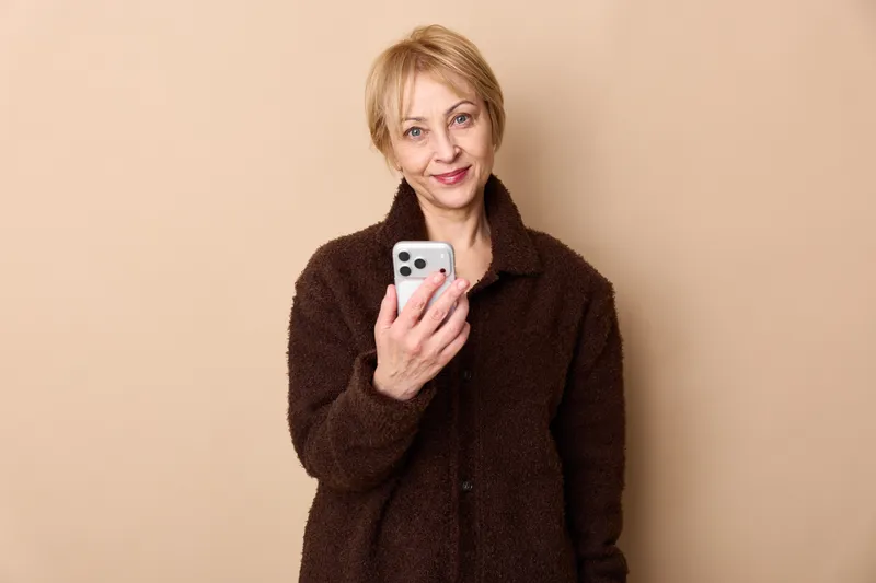 Mature woman holds a smartphone in a portrait indoors on beige background, looking surprised and waiting, showing communication and technology with a dark coat