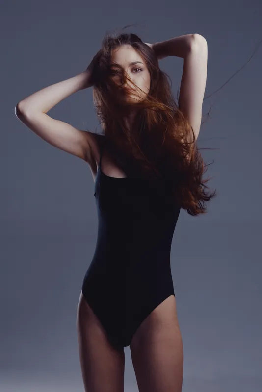Young woman in black bodysuit posing with hands behind head, long hair covering face, moody studio background