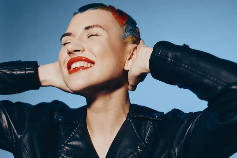 joyful woman with colorful braided hair and bright red lipstick wearing black leather jacket, smiling with closed eyes against dark background, showing happiness and confidence.