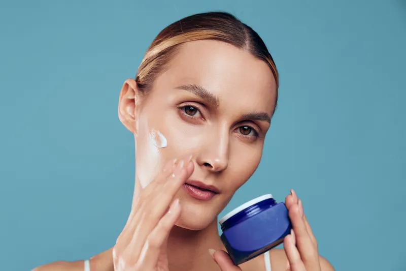 Clear close up of a woman applying moisturizer, showcasing skincare routine, calm expression, soft lighting, smooth skin, blue background, natural glow, fresh complexion