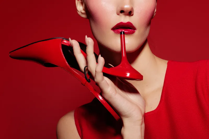 Close-up of woman with red lipstick holding red high heel shoe near lips on red background. Concept of fashion, beauty, style, and elegance with vibrant monochrome color scheme.