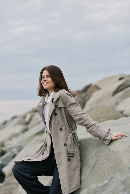Young woman in a grey coat sits on rocks near the sea under a cloudy sky. She smiles calmly, enjoying the peaceful outdoor scene with natural tones and casual fashion style.