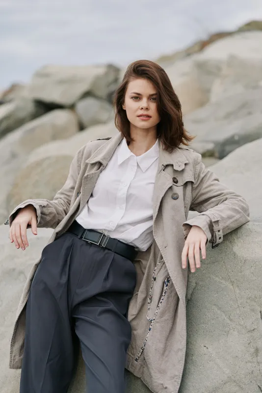 Young woman with brown hair wearing a textured coat and white shirt leans on large rocks outdoors. Natural light highlights her relaxed posture and calm expression. Casual fashion concept.