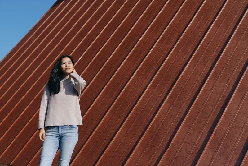 Woman standing against a large diagonal rust wall in outdoor light, wearing a cozy sweater and jeans, confident pose, street style inspired scene with urban backdrop.