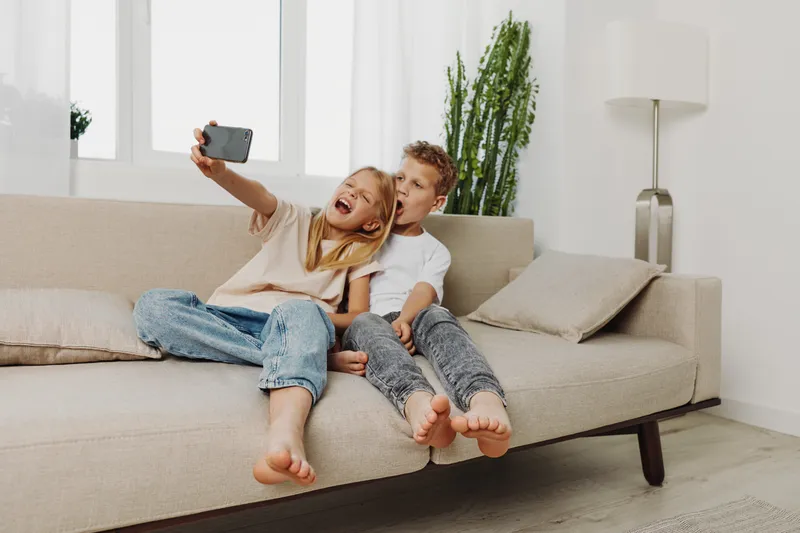 Two cheerful kids sitting barefoot on beige sofa taking selfie with smartphone in bright living room. Concept of family, childhood joy, casual lifestyle, and technology interaction.