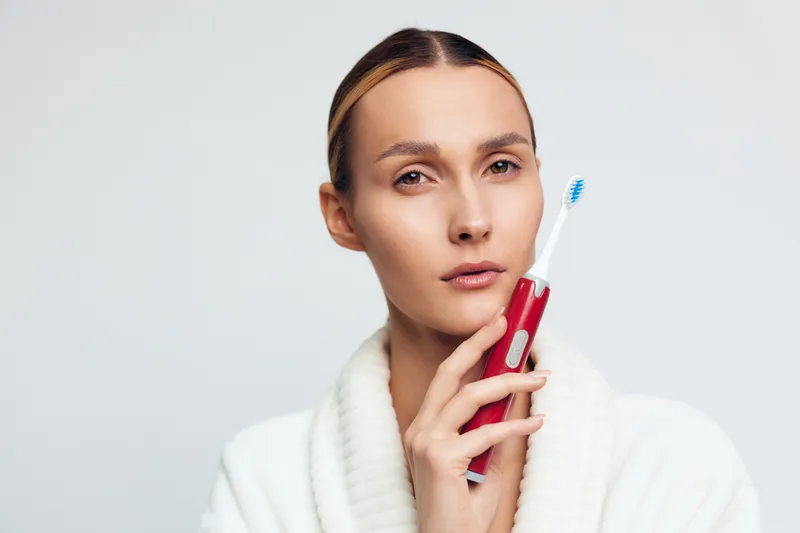 A young woman in a white sweater holds a red electric toothbrush near her face, looking thoughtfully at the camera against a plain white background. Concept for dental care.