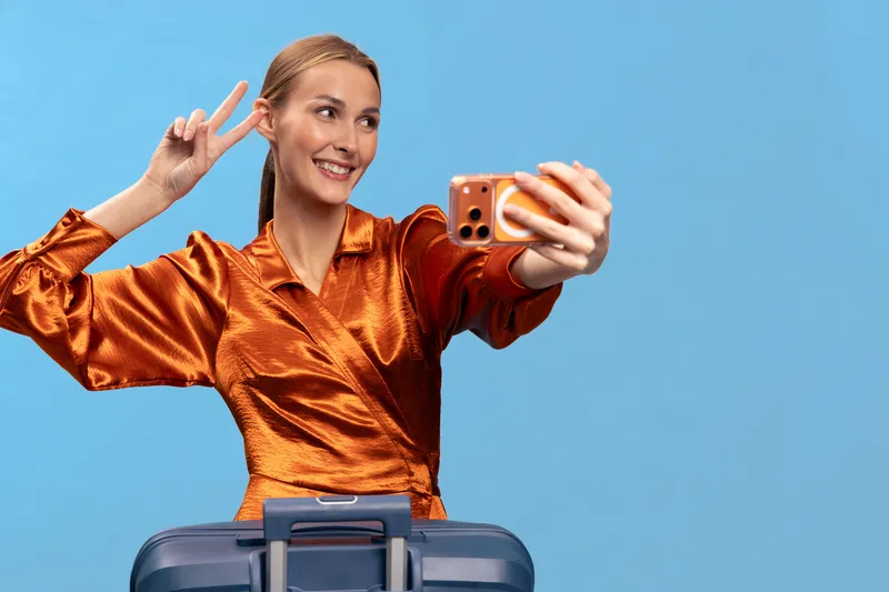 Smiling woman in shiny orange blouse takes selfie with smartphone and shows peace sign. Blue background and suitcase in foreground. Concept of travel, vacation, and joyful moments.