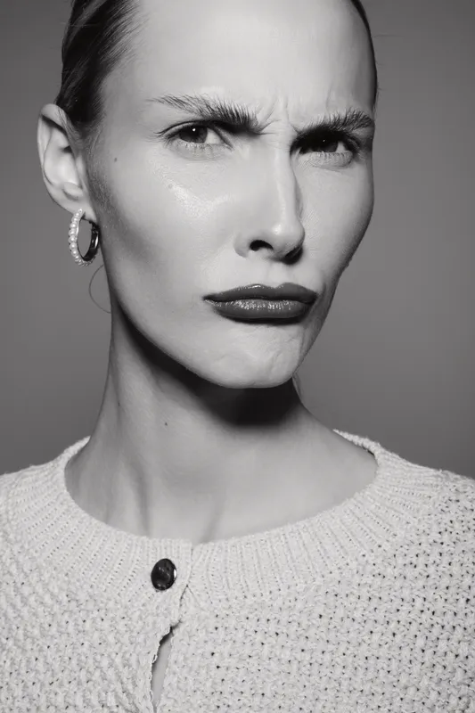 Closeup of a serious white woman with pale skin and earrings frowning in a knitted sweater on gray background.