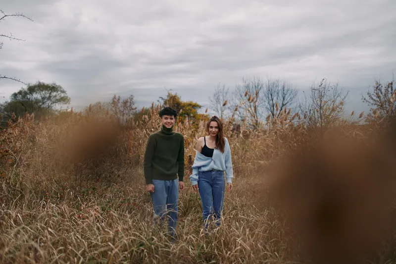 Two young adults walk through tall autumn grass in a candid outdoor portrait, conveying natural connection and a sense of authenticity while capturing accurate everyday moments and subtle expressions.