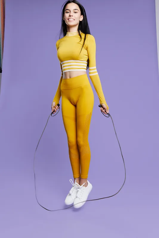 Woman sport fitness jumping rope in yellow activewear against a purple background, demonstrating energy, flexibility, and workout motivation in modern athletic style.