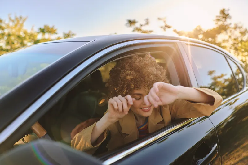 Casual candid shot of a woman driving a car, enjoying warm sunlight, retro film color mood, relaxed lifestyle scene, everyday travel and freedom captured during golden hour.