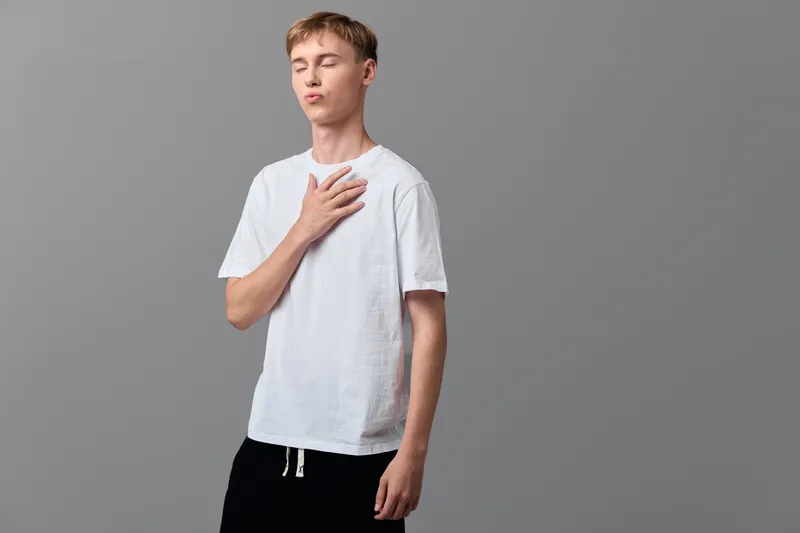 Calm breathing, handonchest, shirt, studio, relaxed pose, focused breath, serene mood, wellness, mindfulness, minimal lighting, backdrop, young male model stands in a neutral studio, simple clothing,