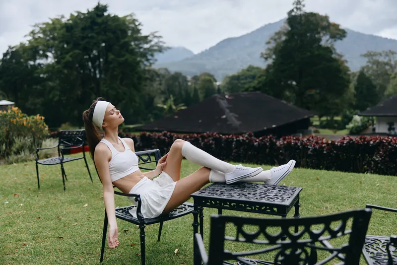 Stylish young woman relaxing in a scenic garden setting, wearing a sporty outfit and exuding a carefree vibe amidst lush greenery and mountains