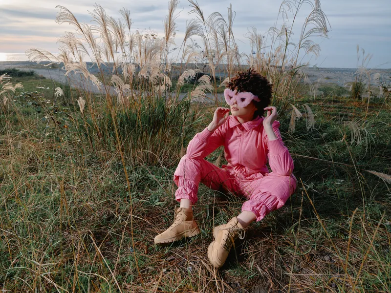 Young person in a vibrant pink jumpsuit sits on tall grass by the sea, boots on, posing for a fashion portrait with natural coastal light and relaxed mood.