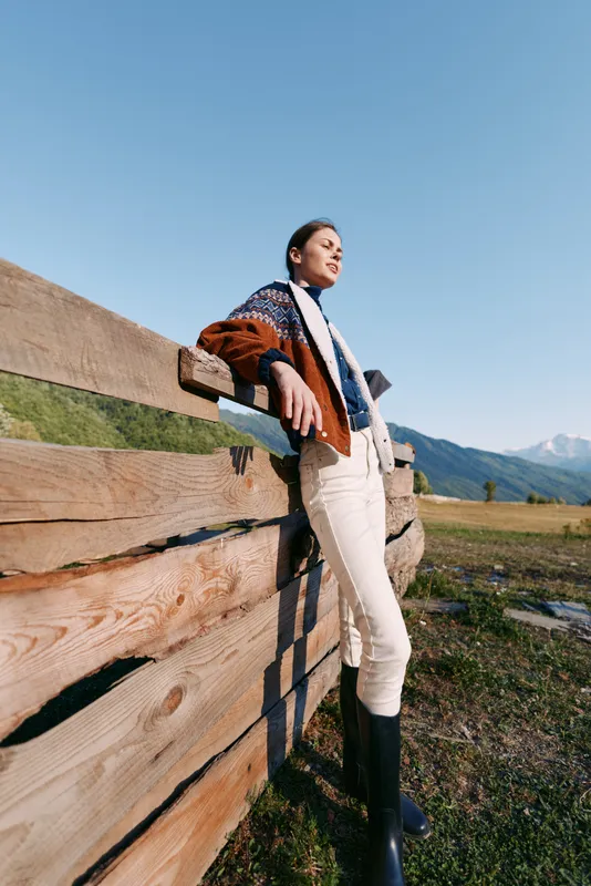 Woman leaning on a wooden fence in the countryside with mountains behind, wearing a cozy sweater, scarf and boots, relaxing outdoors on a sunny clear day in a stylish outfit.