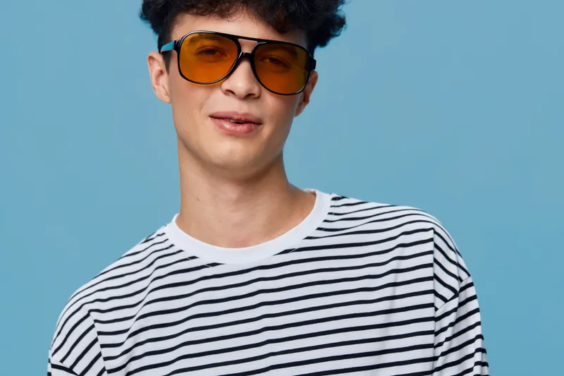 Young man wearing round orange sunglasses and a striped white and black shirt posing confidently against a plain blue background, casual style and modern look.