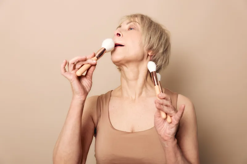 Mature woman with short hair holds two makeup brushes near face, demonstrating joyful expression on beige background. Concept of beauty, self-care, aging, and confidence.