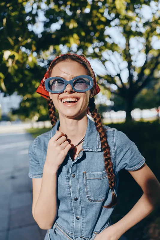 Lifestyle woman in modern boho-western denim street style smiling outdoors, wearing large blue sunglasses and braided hair under natural sunlight, film color effect.