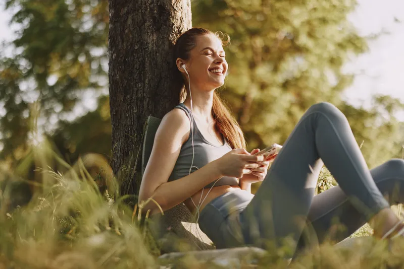Young woman enjoying music while relaxing outdoors in a sunny park, wearing casual sportswear, representing happiness and tranquility in nature