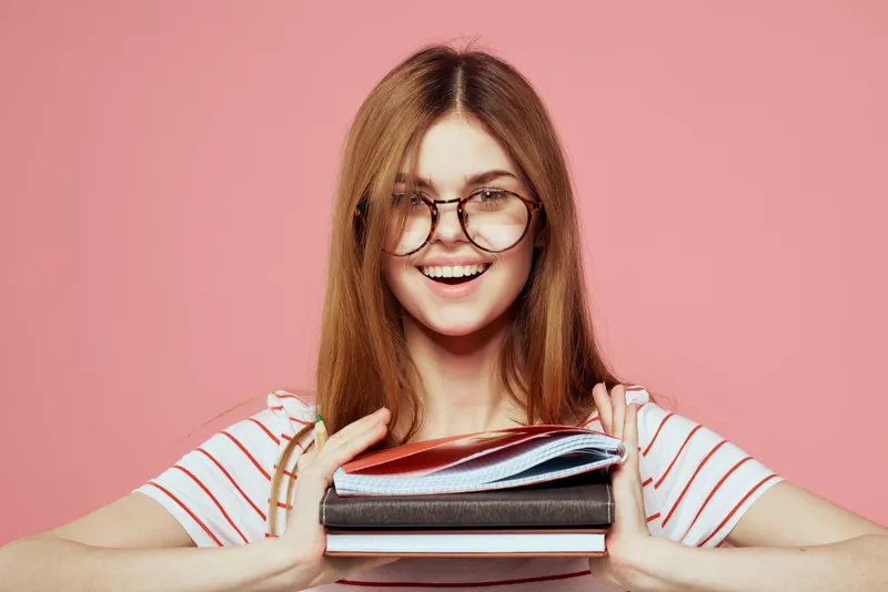 Молодая студентка с книгами на розовом фоне, в очках, крупный план лица