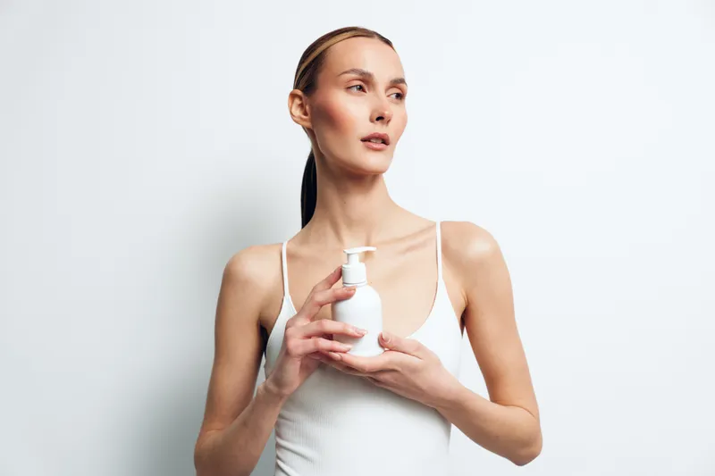 A young woman in a white tank top holds a white pump bottle against a plain white background. She looks to the side with a calm expression, concept for skincare or beauty product.