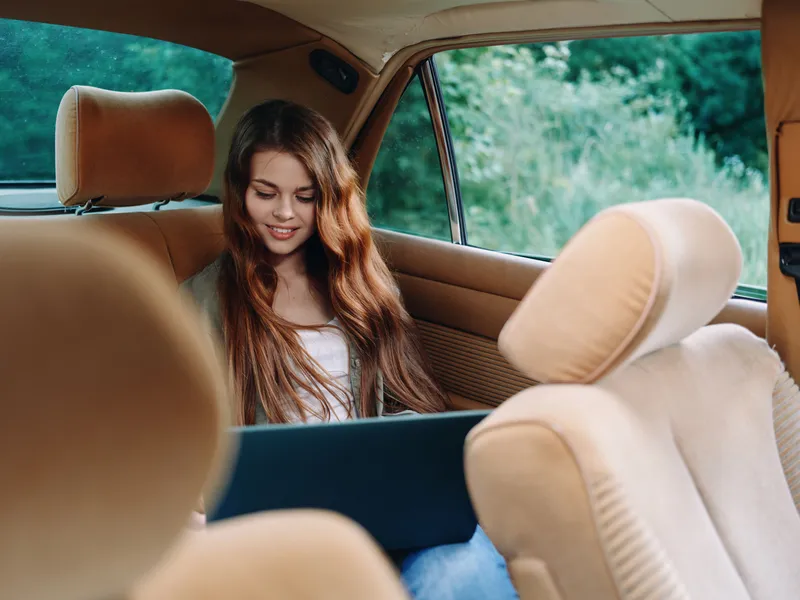 Young woman working on a laptop in a vintage car during a sunny day, dressed casually with long hair, surrounded by nature in a relaxed atmosphere.